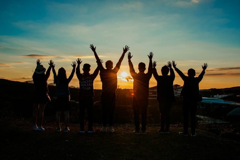Photo by Min An A diverse group of friends raises their arms in celebration against a vibrant sunset backdrop.